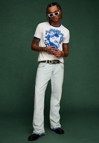 Bela majica s modrim obrubom in grafičnim dizajnom, svetlo modri naravnost krojeni traper hlače, črn pas in masivni črni čevlji. Dodatki: bele sončna očala.