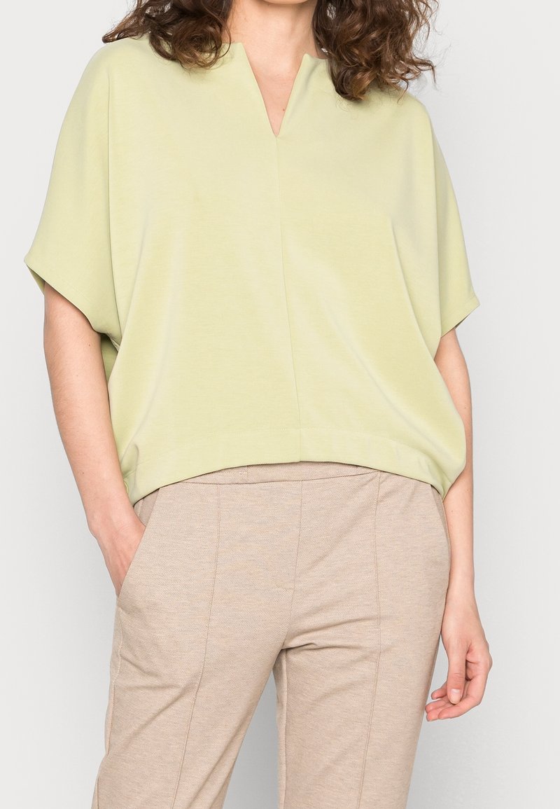 Femme portant un haut ample à manches courtes de couleur vert clair avec un col en V et un pantalon beige ajusté, les mains dans les poches.
