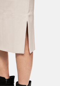 Helle beige Skirt mit einem seitlichen Schlitz, aus weichem Stoff gefertigt. Ein Paar schwarze Stiefeletten sind am unteren Rand sichtbar und verstärken den Kontrast des Outfits.
