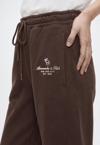 Близък план на кафяви спортни панталони с връзка и бяло лого на Abercrombie & Fitch, бродирано над джоба, ръка, поставена на бедрото.