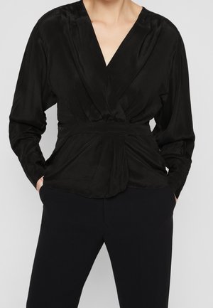 Femme portant un chemisier cache-cœur noir à manches longues avec un profond décolleté en V et un pantalon noir, mains partiellement dans les poches, sur un fond gris clair.