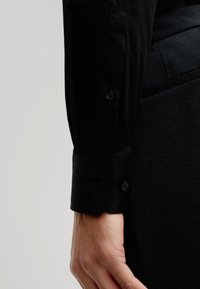 Zwarte shirtmanchet met twee knopen, slanke textuur en geborduurd design op de mouw, gecombineerd met een donker patroonbroek.