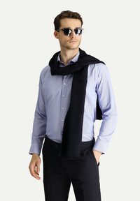 Sciarpa nera drappeggiata su una camicia a righe blu chiaro con bottoni. Modella che indossa occhiali da sole neri, posando su uno sfondo neutro.