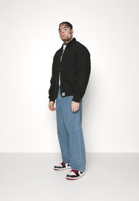 Black zip-up bomber jacket with ribbed cuffs, paired with light blue loose-fitting jeans and white sneaker-style shoes with red accents.