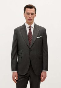 Hombre con traje gris oscuro, camisa blanca, corbata roja de patrones y pañuelo blanco en el bolsillo, de pie frente a un fondo blanco liso.