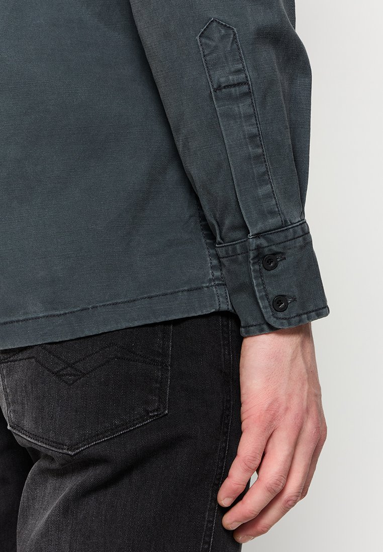 Dunkelgraues Hemd mit spitzen Manschetten, ausgestattet mit zwei schwarzen Knöpfen und einer sauberen, glatten Textur. Schwarze Jeans sind am unteren Ende sichtbar.