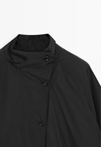 Veste isolante noire avec un col montant, dotée de deux boutons et d'une texture lisse. Conçue avec des coutures angulaires et un style minimaliste.