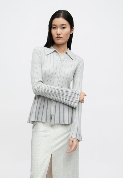 Svetlosivý rebrovaný kardigán so špicatým golierom, zapínaním na gombíky a dlhými, rozšírenými rukávmi. Má striedavé vertikálne pruhy v tmavšej šedej farbe.