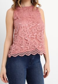 Krajkový top bez rukávů v jemné růžové barvě, s květinovým vzorem, vroubkovaným lemem a hladkou texturou, kombinovaný s tmavě modrými džínami.