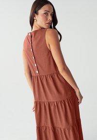 Rostfarbenes ärmelloses Kleid mit einem gestuften Design, das einen Knopfverschluss auf der Rückseite und einen weichen, strukturierten Stoff aufweist.
