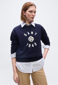 Νεαρή γυναίκα που φοράει μπλε φούτερ GANT πάνω από ριγέ πουκάμισο με κουμπιά και μπεζ παντελόνι, ποζάροντας με το ένα χέρι στην τσέπη μπροστά σε λευκό φόντο.