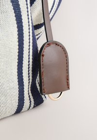Gros plan d’un sac en tissu rayé bleu marine et blanc avec une extrémité de sangle en cuir marron et un petit anneau métallique doré.