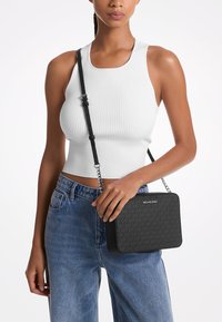 Sieviete, tērpta baltajā ribotajā bez piedurknēm topā un zilos džinsos, tur melnu Michael Kors crossbody somu ar rakstainu logo.