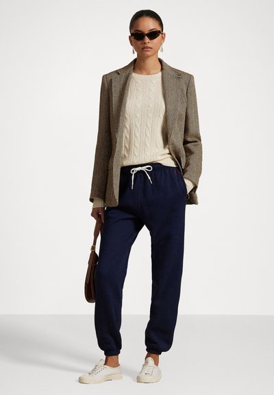 Brown herringbone blazer over a cream cable-knit sweater, paired with navy joggers and white sneakers. Accessorized with black sunglasses and a brown bag.