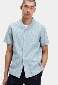 Man in een lichtblauw, kort mouw, gestructureerd overhemd met een wit T-shirt eronder, staand met één hand in zijn zak.