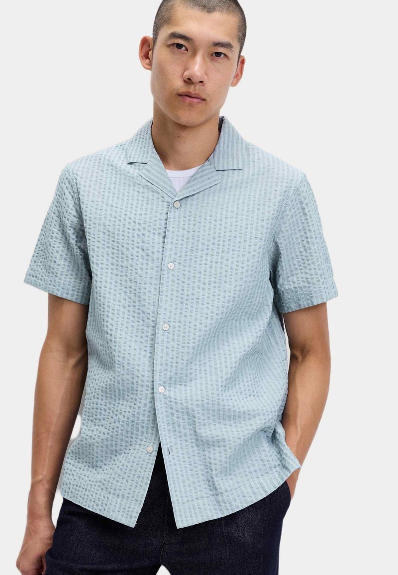 Man in een lichtblauw, kort mouw, gestructureerd overhemd met een wit T-shirt eronder, staand met één hand in zijn zak.