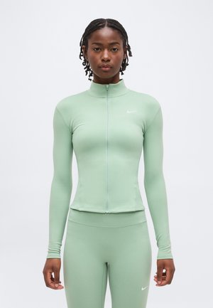 Femme aux cheveux courts tressés portant une veste de sport zippée vert clair et des leggings assortis, debout devant un fond blanc uni.