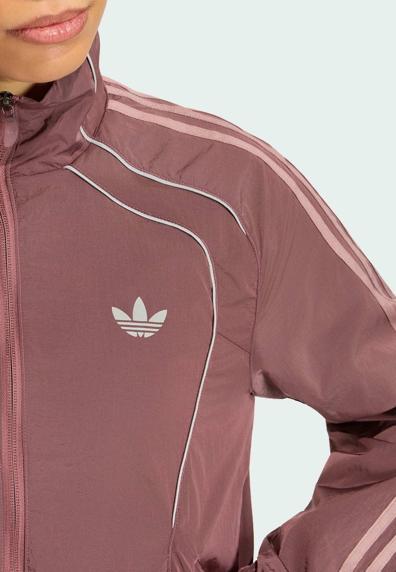 Femme en veste Adidas mauve avec zip et logo sur la poitrine, avec des bandes rose clair sur les épaules et les manches.