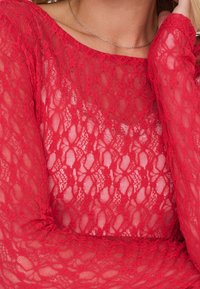 Femme portant un top en dentelle rouge à manches longues et un fin collier argenté, avec une partie de son visage et de son bras visible.