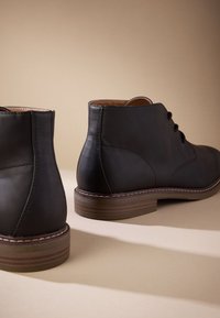 Čierne kožené členkové čižmy s textúrou, šnurovací dizajn a hnedý stohovaný podpätok. Výrazné prešívanie pozdĺž okrajov.