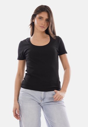 Giovane donna con capelli castani lisci che indossa una maglietta nera a maniche corte e jeans azzurri, in piedi davanti a uno sfondo semplice.