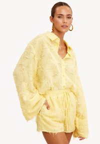 Chemise jaune texturée oversize avec col et boutons, associée à un short assorti. Présente un détail doux ressemblant à des franges et une taille élastique.