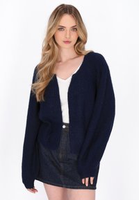 Jonge vrouw met lang blond haar, gekleed in een marineblauwe cardigan, witte top en een donkere denim rok, staand tegen een witte achtergrond.