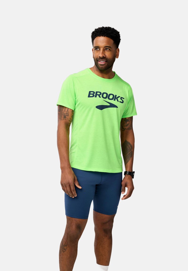 Camiseta de running verde neón con el logo de "BROOKS" en navy, de manga corta, combinada con pantalones cortos navy. Tejido suave y ligero, ajuste atlético.