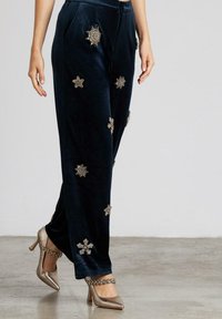 Pantalones de terciopelo azul marino con adornos florales en plata y oro, con un corte relajado y dos bolsillos laterales, combinados con tacones metálicos.