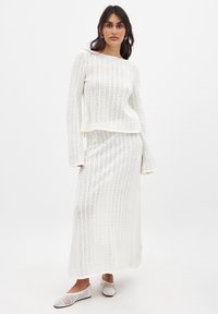 Valge kootud kahest osast koosnev komplekt, mis sisaldab pikka varrukatoppi ja maksi seelikut, mõlemad tekstuurse mustriga ja vabalt voolava lõikega.