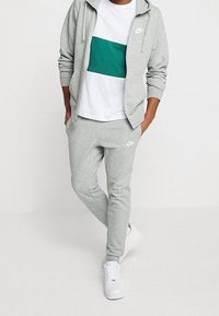 Man i ljusgrå Nike-hoodie och träningsbyxor med vita skor och vit T-shirt med grön rand, står med händerna i fickorna.