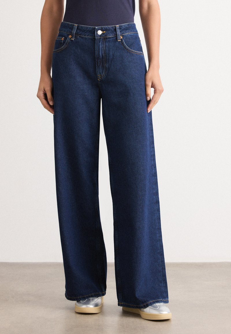 Jean en denim à jambes larges, de couleur bleu foncé, avec des coutures orange, des poches avant et une fermeture en métal. Texture lisse et coupe décontractée.