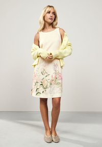 Robe en lin imprimée floral en jaune pâle avec un col rond, associée à un cardigan jaune doux. Comprend des fentes latérales et une coupe décontractée.
