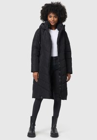 Long manteau rembourré noir avec capuche, présentant une fermeture éclair et un motif de matelassage diagonal, associé à un jean foncé et des bottes noires.