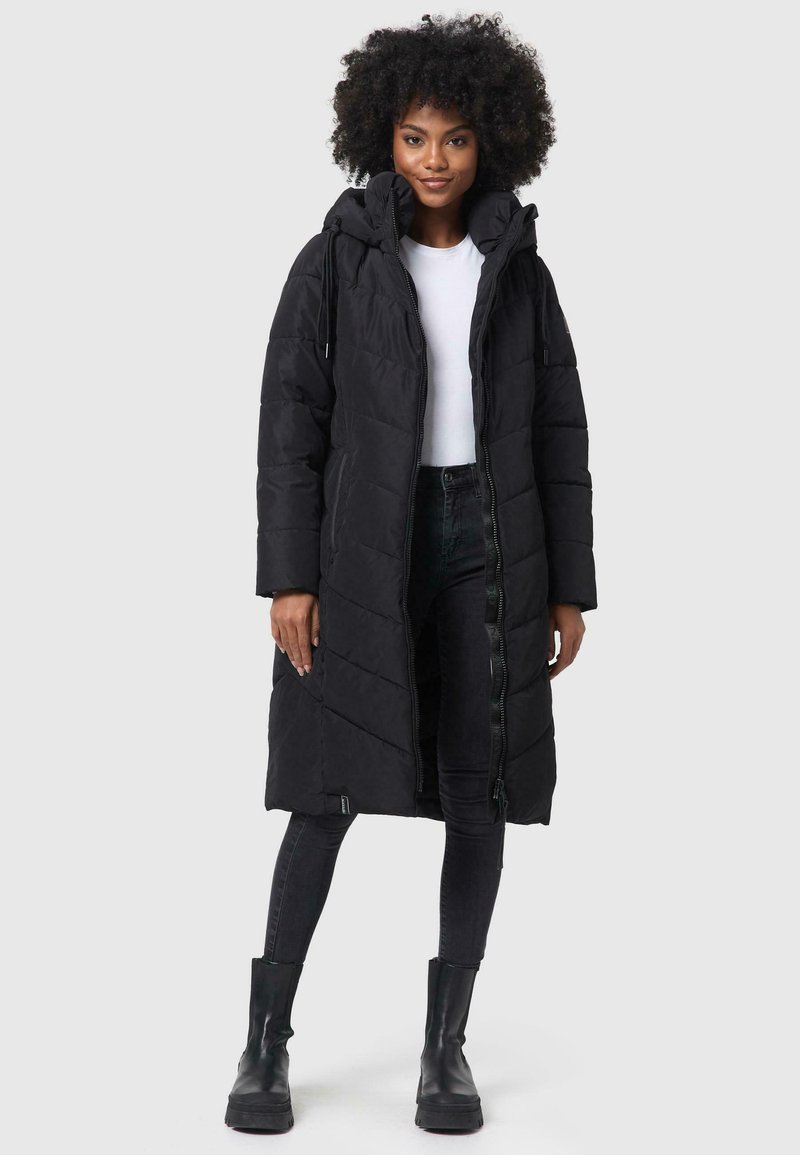 Long manteau rembourré noir avec capuche, présentant une fermeture éclair et un motif de matelassage diagonal, associé à un jean foncé et des bottes noires.