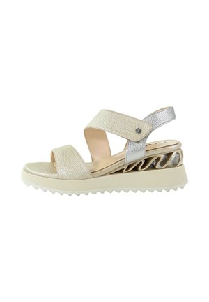 Sandał na koturnie z beżowymi skórzanymi paskami i srebrnym paskiem z tyłu, z unikalnym falistym metalicznym wzorem na podeszwie oraz białą, rowkowaną podeszwą zewnętrzną.