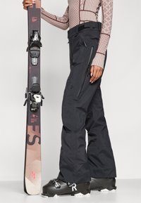 Black ski pants with multiple pockets, adjustable waistband, and waterproof material, paired with black ski boots and a patterned ski.