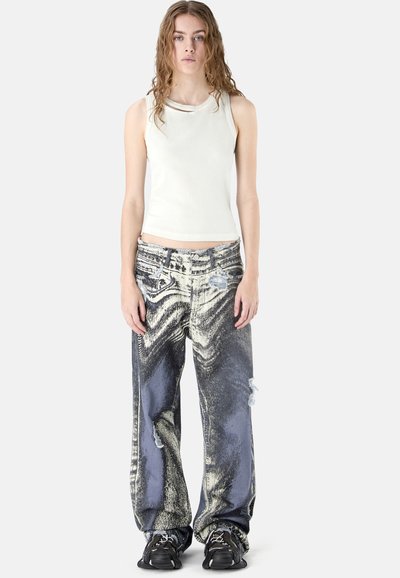 Jeune femme aux cheveux ondulés portant un haut blanc sans manches, un jean ample délavé avec des déchirures, et des baskets noires, debout devant un fond blanc.