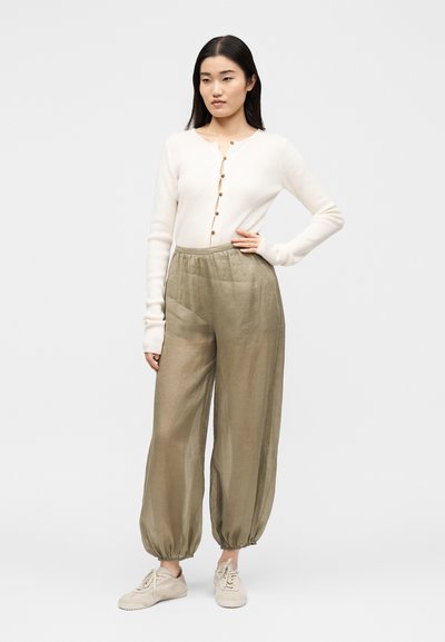 Femme debout, main sur la hanche, portant un cardigan blanc boutonné, un pantalon ample vert olive avec des poignets élastiques et des baskets beige.