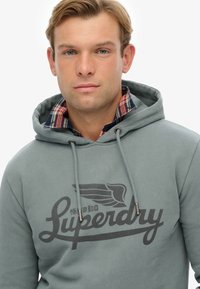 Grijze hoodie met een logo aan de voorkant en een trekkoordsluiting, gedragen over een geruit flanellen overhemd met blauwe en rode patronen.