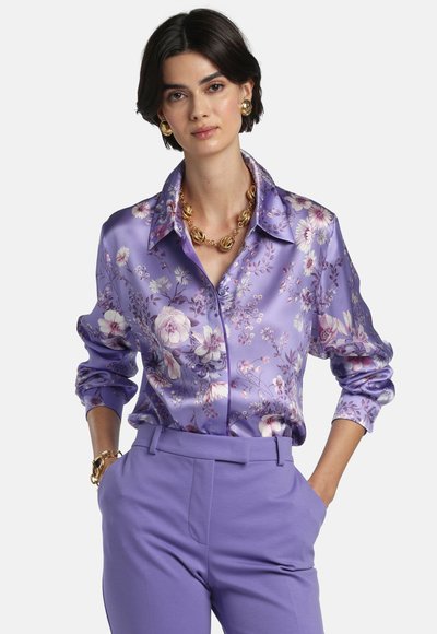 Woman wearing a purple floral satin blouse with long sleeves and purple high-waist pants, accessorized with gold jewelry.