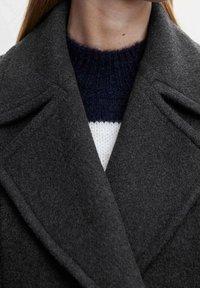 Charcoalgrijs wollen jas met een opstaande kraag, voorzien van een subtiele textuur en een getailleerd ontwerp. Draag het over een donkerblauw en wit gestreept trui.