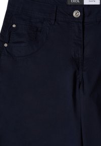 Pantalon bleu marine foncé en gros plan montrant la poche avant, la fermeture par bouton, la boucle de ceinture et une étiquette partielle de la marque indiquant Cecil Pants.