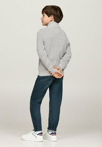 Maglione grigio a maglia con collo alto, abbinato a jeans in denim blu scuro e sneakers bianche. Il bambino sta in piedi con le mani dietro la schiena.