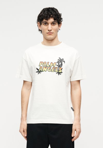 Νέος άνδρας με σγουρά σκούρα μαλλιά που φοράει λευκό T-shirt με τα γράμματα "PALM ANGELS" και γραφικά με φοίνικες, στέκεται μπροστά σε ένα απλό φόντο.
