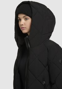 Chaqueta de plumas acolchada negra con una gran capucha, que presenta costuras diagonales, cierre frontal con cremallera y un bolsillo lateral con cremallera.