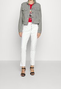 Černobílý kostkovaný zkrácený kabát přes červené tričko s grafikou, doplněný bílými kalhotami s vysokým pasem a černými sandály na podpatku.