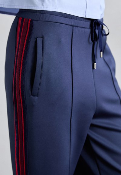 Pantalones deportivos azul marino con rayas rojas, que cuentan con bolsillos laterales y una cintura elástica con cordones. Textura de tela suave y lisa.
