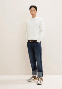 Pull léger blanc, jean en denim noir avec ourlets retroussés, et baskets multicolores avec un mélange de textures et de motifs.