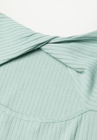 Hellgrünes, geripptes Top mit einer gedrehten Detail an der Rückseite, das eine weiche Textur und einen runden Halsausschnitt aufweist. Sichtbare Nähte setzen Akzente.